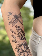 Tattoo of a frog, dragonfly, tadpoles, and lotus flower on a person's arm with a blurred natural background.