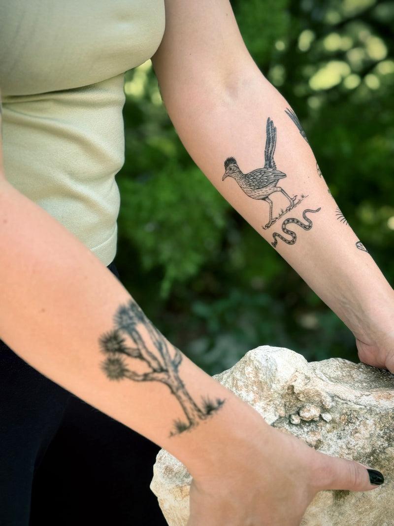 Person with temporary tattoos on arms and legs, standing outdoors with greenery in the background