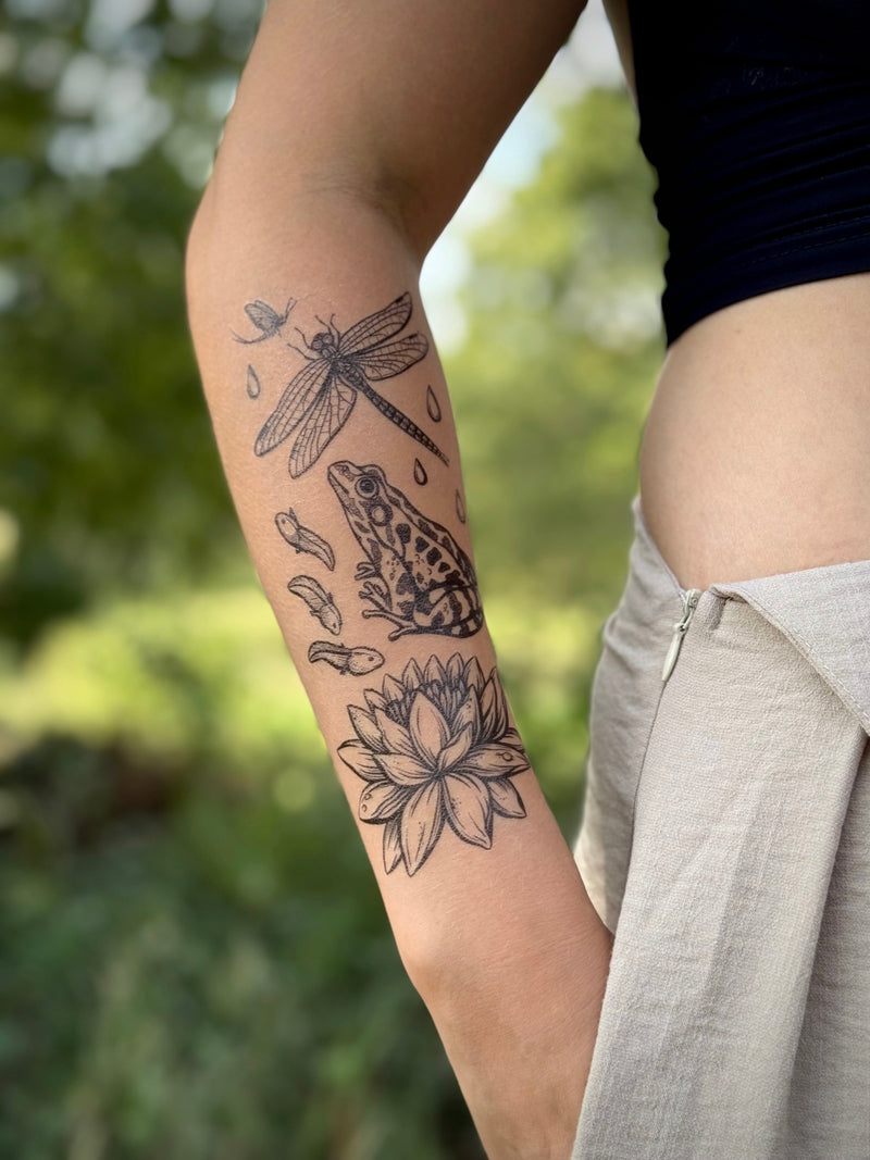 Arm with nature-themed temporary tattoos of a frog, lotus flower, and dragonfly against a blurred natural background.