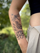 Arm with nature-themed tattoos of a frog, lotus flower, and dragonfly against a blurred natural background.