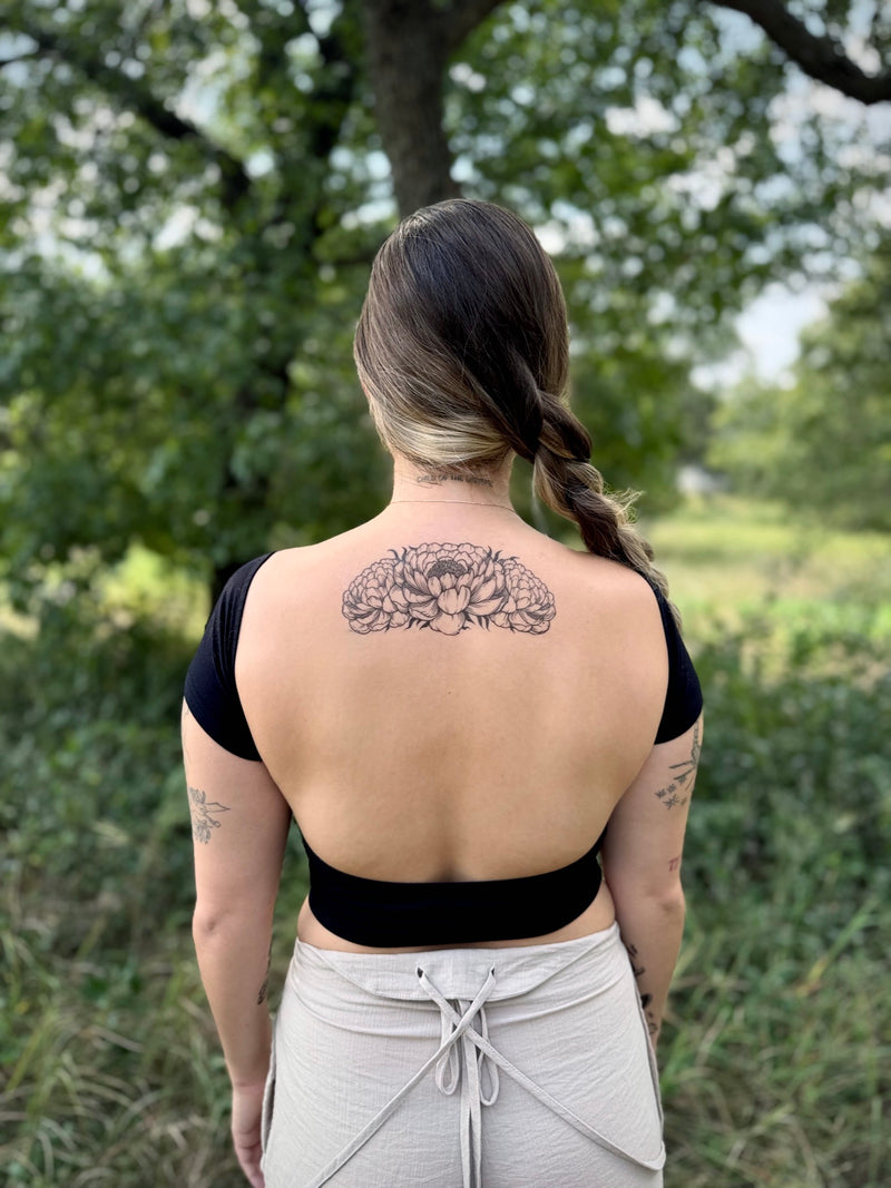 Person with a peony temporary tattoo on their back standing in a natural setting with trees and greenery.
