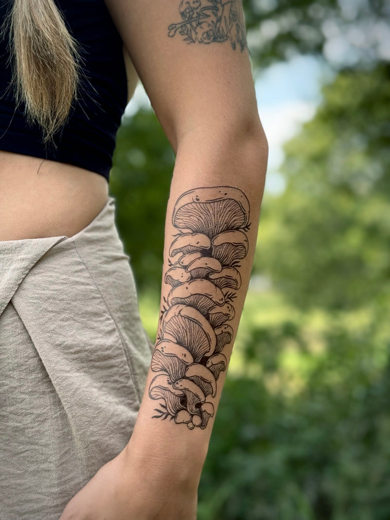 Person with a detailed mushroom temporary tattoo on their arm, standing outdoors with greenery in the background.