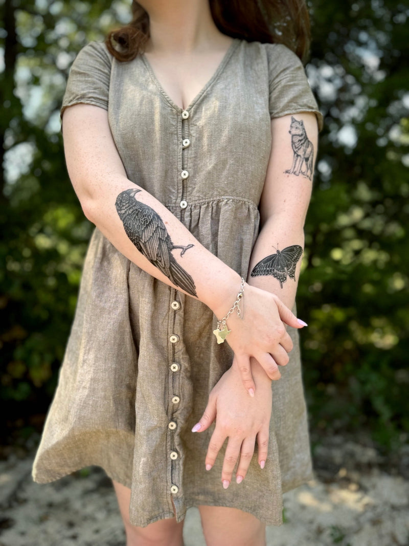 Woman wearing a brown dress with visible temporary tattoos on her arms against a blurred natural background