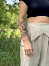 Person with tattoos on their arm standing outdoors with greenery in the background