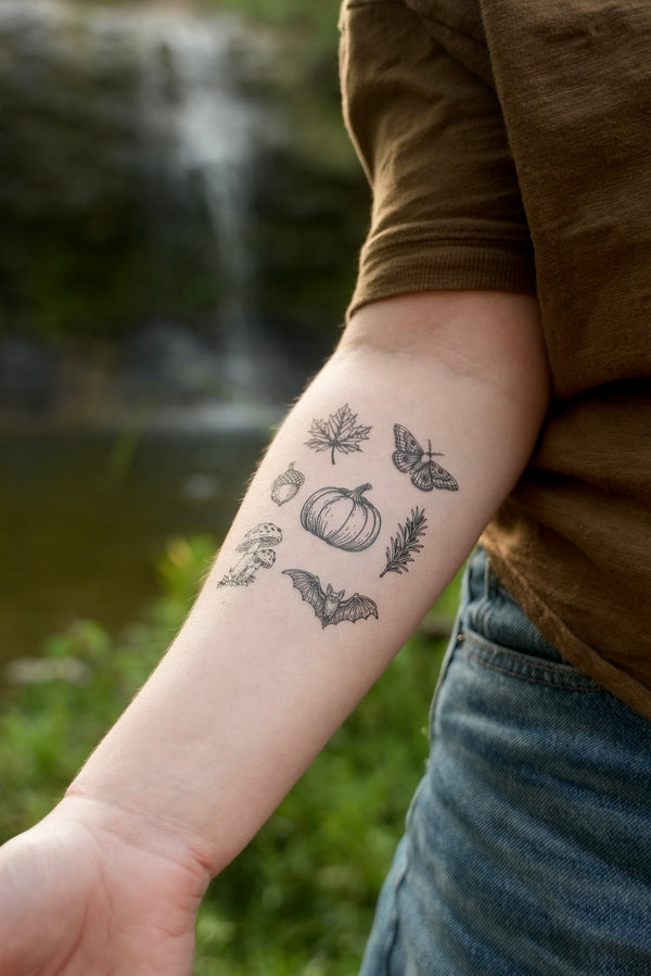 A temporary tattoo applied to a person's forearm, featuring black line art illustrations of autumn and Halloween motifs including a pumpkin, maple leaf, moth, acorn, bat, rosemary, and mushroom.