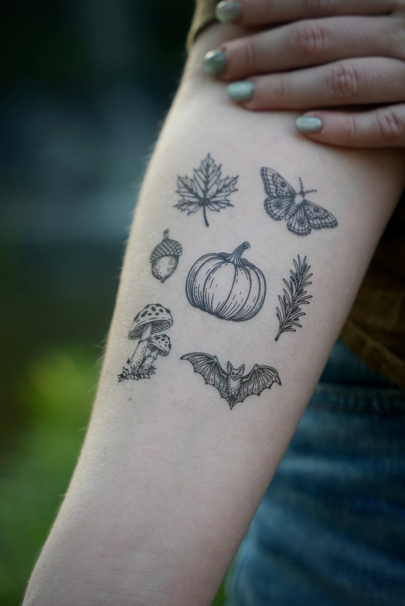 A temporary tattoo applied to a person's forearm, featuring black line art illustrations of autumn and Halloween motifs including a pumpkin, maple leaf, moth, acorn, bat, rosemary, and mushroom.