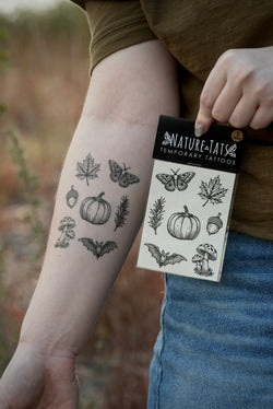 A person outdoors holds a package of Nature's Tattoos temporary tattoos next to their forearm, which shows several black ink designs including a pumpkin, maple leaf, moth, acorn, rosemary and mushroom.