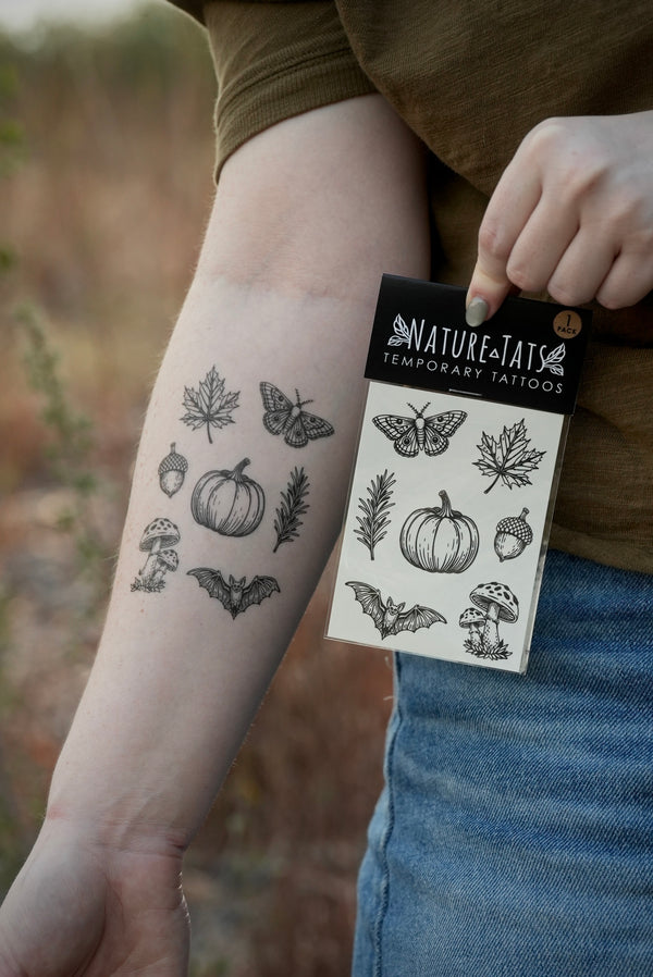 A person outdoors holds a package of Nature's Tattoos temporary tattoos next to their forearm, which shows several black ink designs including a pumpkin, maple leaf, moth, acorn, rosemary and mushroom.