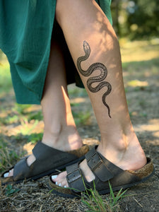 Person wearing black sandals with a snake tattoo on their leg, standing on grass.