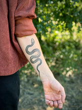 Person with a snake tattoo on their arm against a blurred green outdoor background
