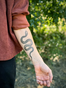 Person with a snake tattoo on their arm against a blurred green outdoor background