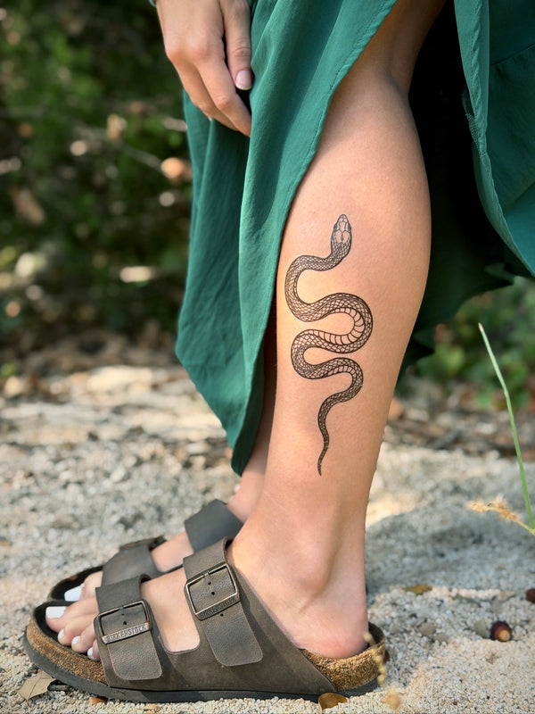Person with a snake temporary tattoo on their leg, wearing sandals and a green garment, standing on sandy ground with blurred greenery in the background.