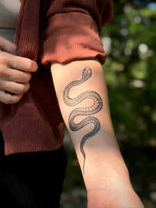 Person wearing a brown sweater with a snake tattoo on their arm, standing outdoors.