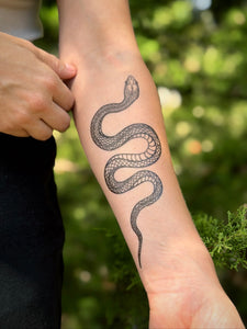 Person with a snake tattoo on their arm, standing outdoors with greenery in the background