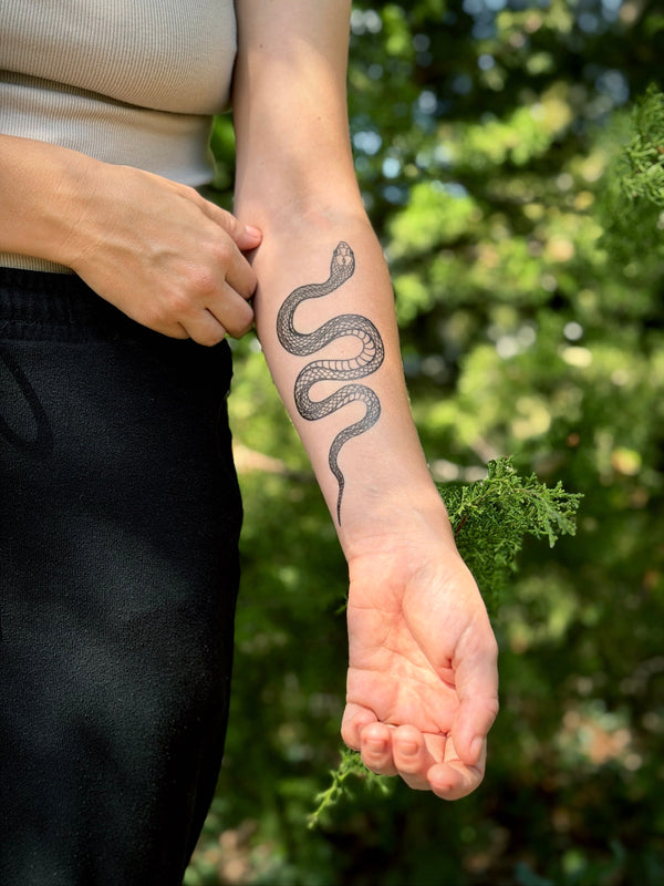 Person with a snake temporary tattoo on their arm standing outdoors with greenery in the background 