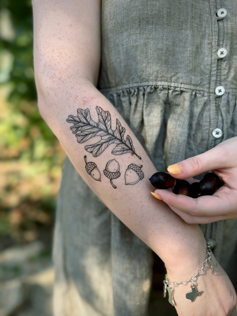 Person wearing a sleeve with leaf and acorn temporary tattoos, holding dark berries outdoors.