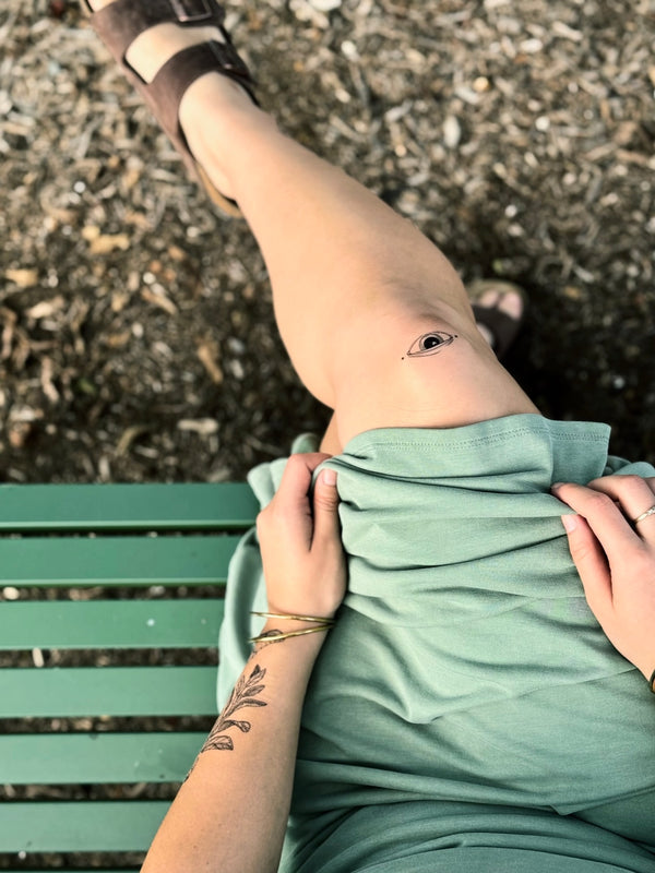 Person sitting on a green bench with a green dress, wearing brown sandals on a textured ground with temporary tattoos on knee and wrist 