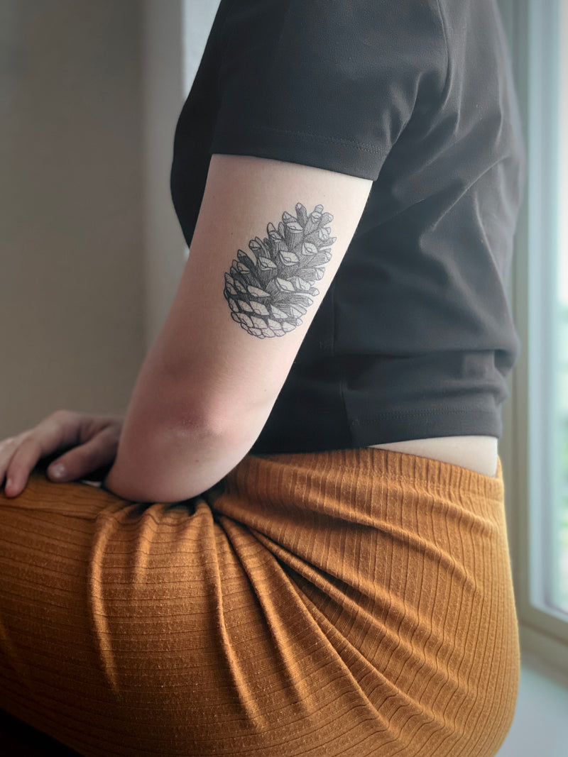 Person wearing a black shirt with a pine cone temporary tattoo on their arm, sitting on a brown chair.
