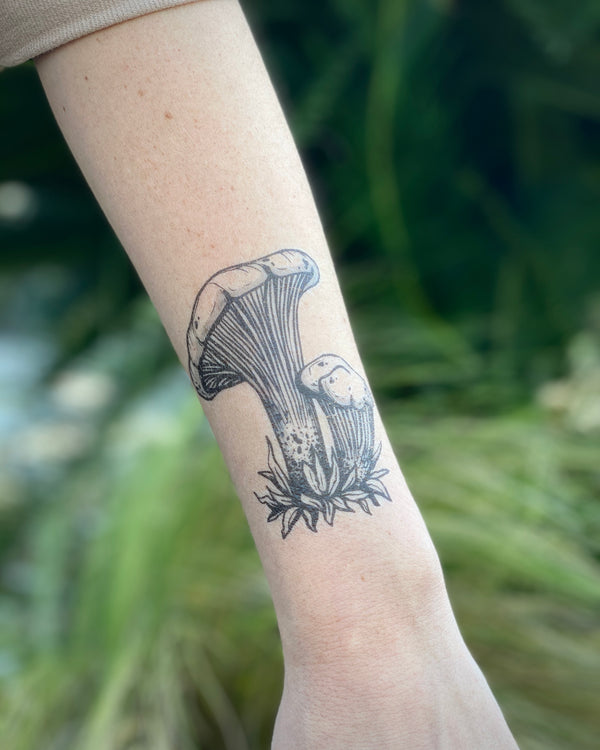 Temporary tattoo of a chanterelle mushroom on a person's wrist, featuring black line art with no color fill, set against a blurred green background.