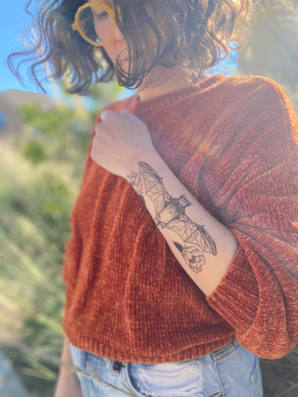 free-tailed bat temporary tattoo on a persons arm wearing an orange sweater and blue jeans in a blurred nature background 