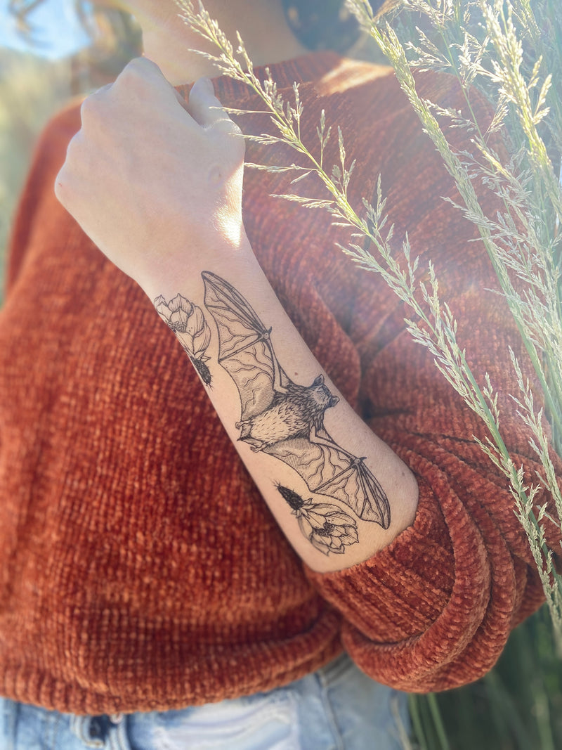 free-tailed bat temporary tattoo on persons arm wearing an orange sweater and blue jeans with a leafy blurred background 