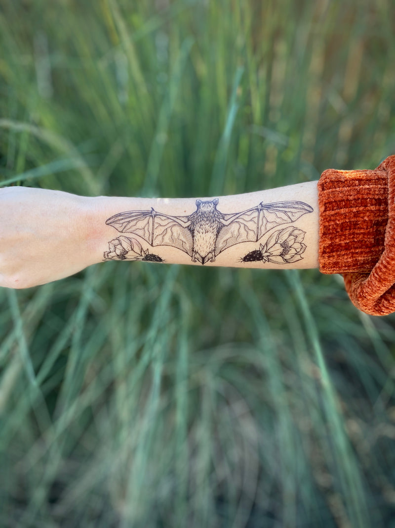 free-tailed bat temporary tattoo on persons arm wearing orange sweater with a wispy green nature background 