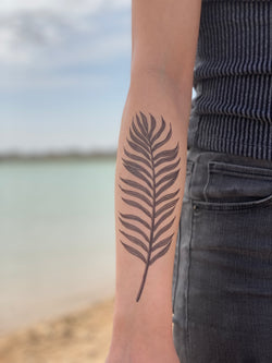 A temporary tattoo of a palm leaf on a person's forearm, displayed against a beach backdrop.