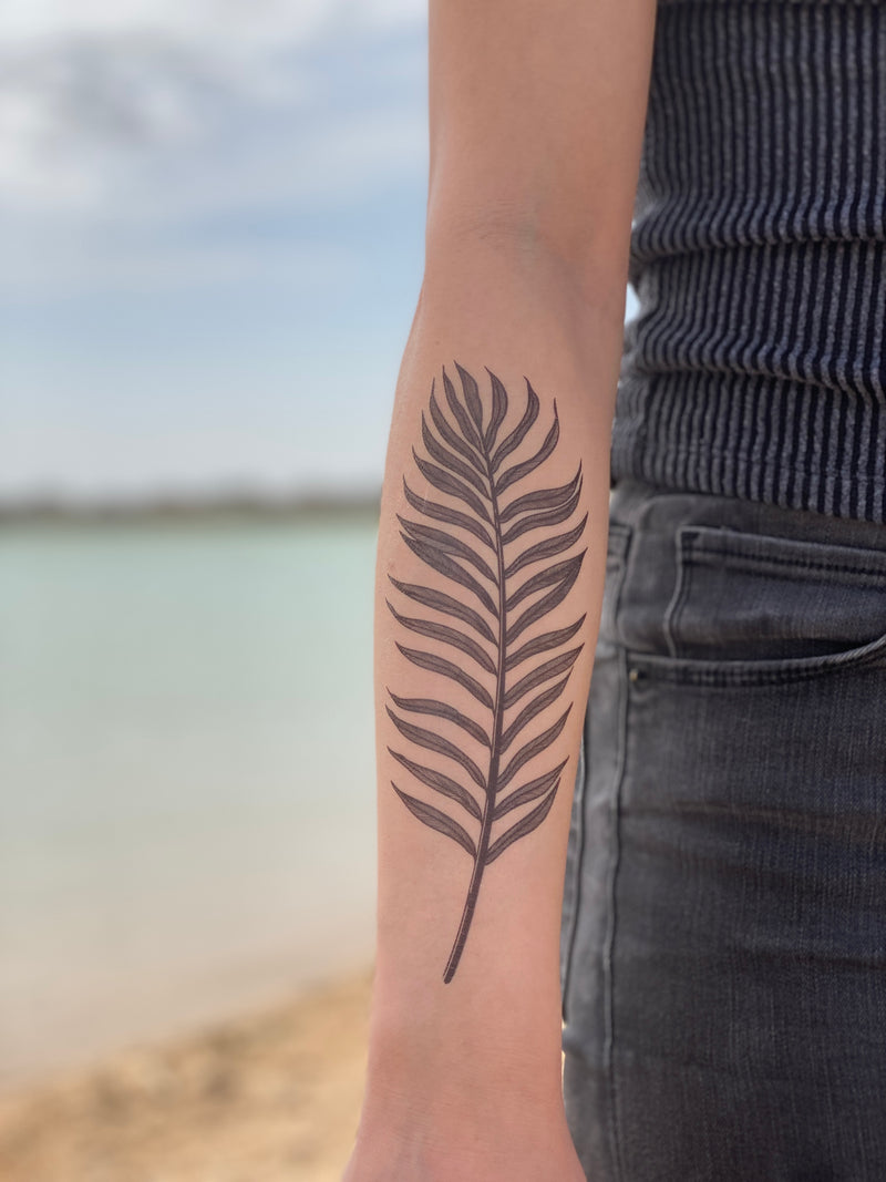A temporary tattoo of a palm leaf on a person's forearm, displayed against a beach backdrop.