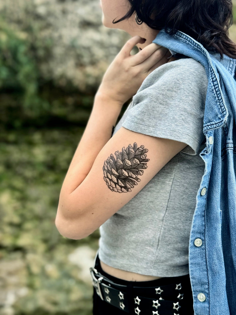 Person with a pine cone temporary tattoo on their arm, wearing a denim jacket and gray crop top, standing against a natural background.