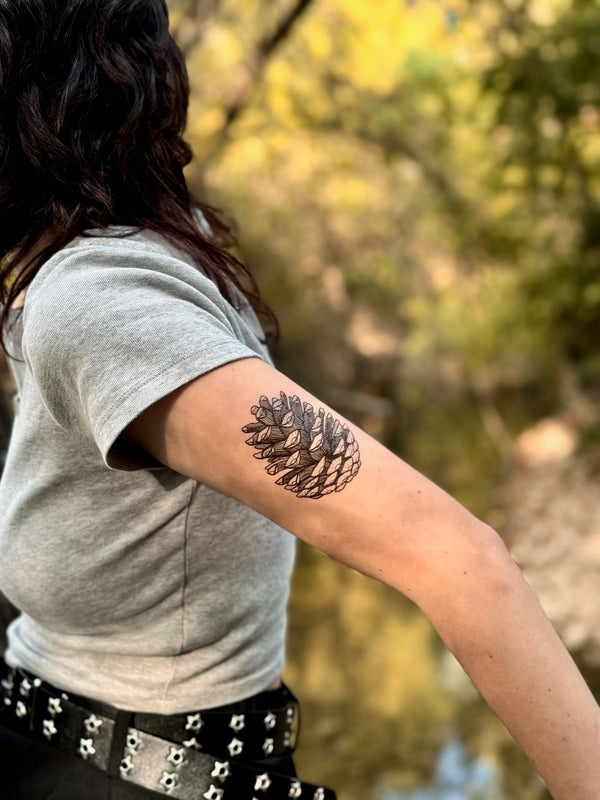 Person with a pinecone temporary tattoo on their arm standing outdoors with trees in the background