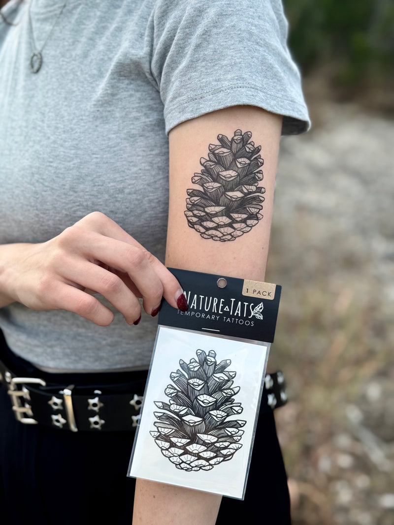 Person with a temporary tattoo of a pine cone on their arm, holding a package of Nature-Tats temporary tattoos.