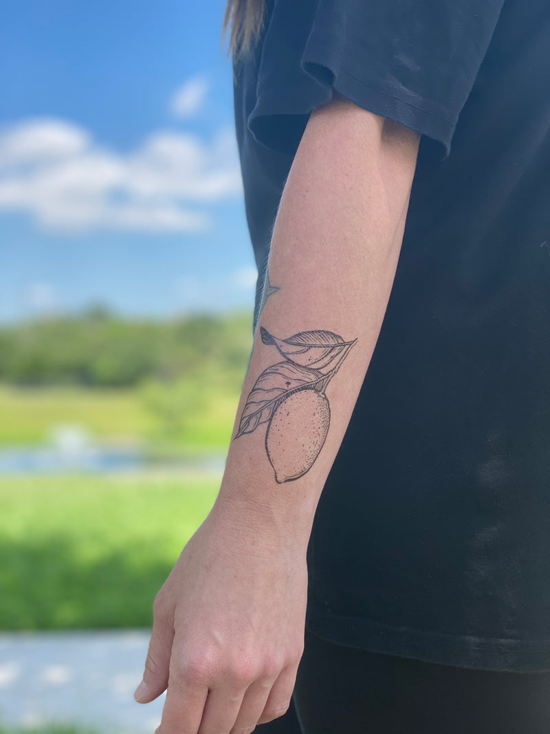 lemon with leaves temporary tattoo on persons wrist wearing black in a blurred nature background 