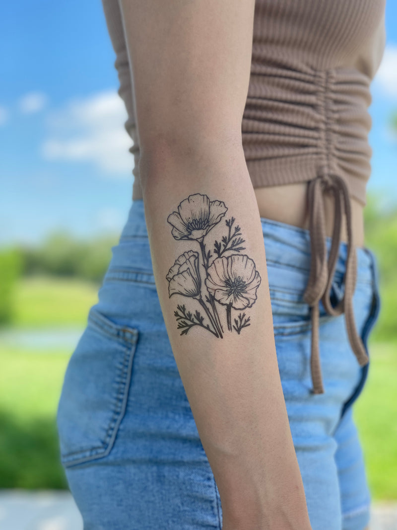 golden poppy temporary tattoo on a persons arm wearing a brown shirt and blue jeans in a blurred nature background 