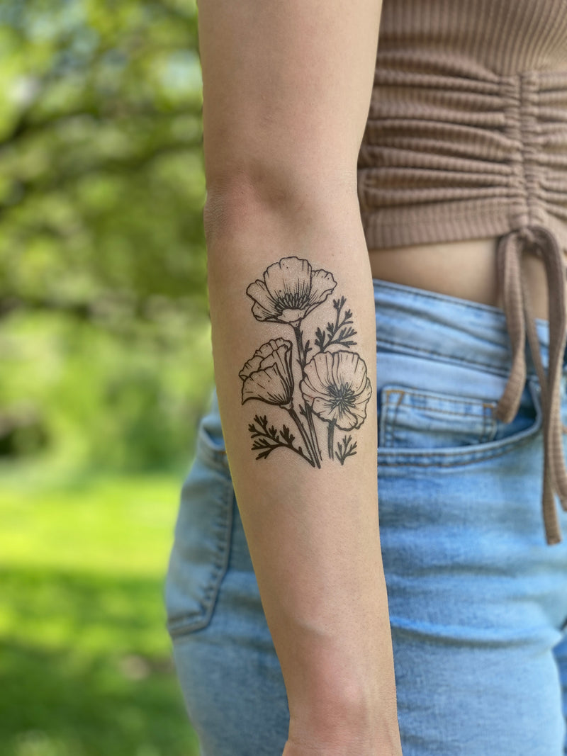golden poppy temporary tattoo on a persons arm wearing a brown shirt and blue jeans in a green blurred nature background 
