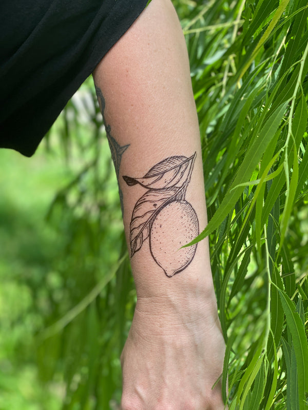 lemon with leaves temporary tattoo on persons wrist wearing black in a green nature background 