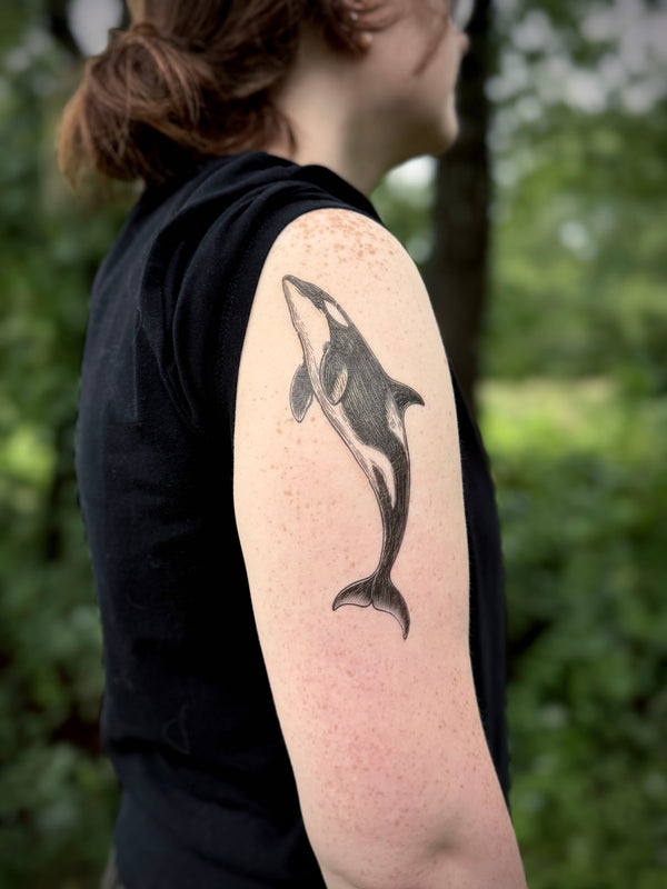 Person with a tattoo of an orca whale on their arm, standing outdoors with greenery in the background.