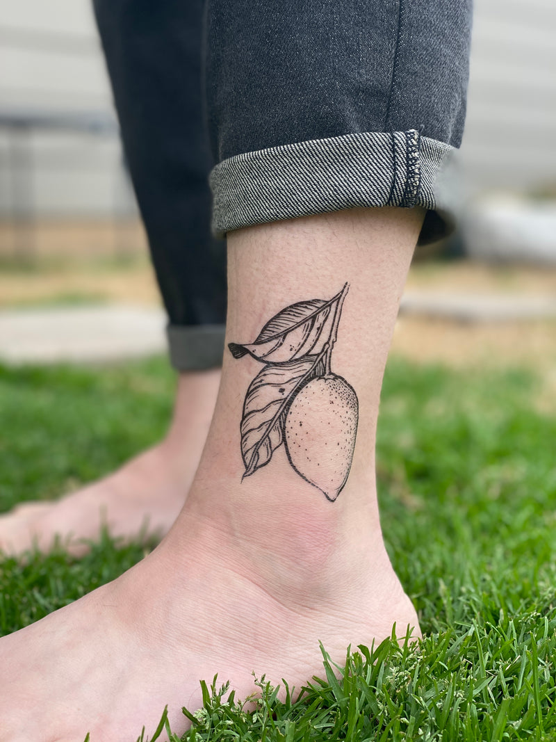 lemon with leaves temporary tattoo on persons ankle wearing black jeans with a grass background 