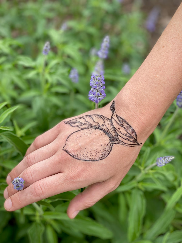 A temporary tattoo of a lemon with leaves on a person's hand, displayed against a background of purple flowers.