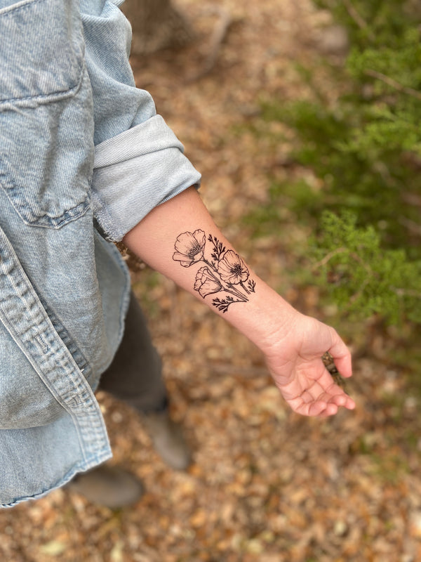 golden poppy temporary tattoo on person arm wearing blue jean clothes in a blurred wooded background 
