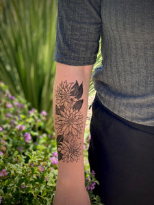 person wearing black ink temporary tattoo of 3 clustered dahlia flowers on forearm with flowers in the background