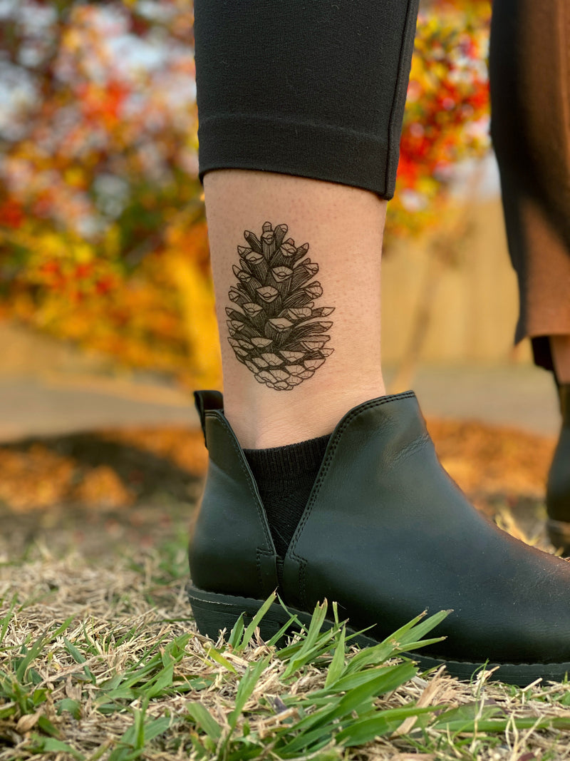 Person wearing black shoes with a pine cone temporary tattoo on a blurred outdoor background