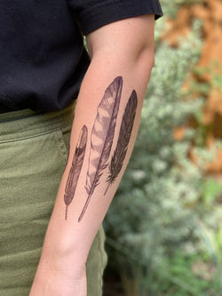 A person's forearm featuring a temporary tattoo with three different feather designs.