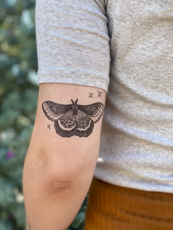 Temporary tattoo of a night moth with details and sparkles on a person's forearm wearing white and orange with a blurred nature background 