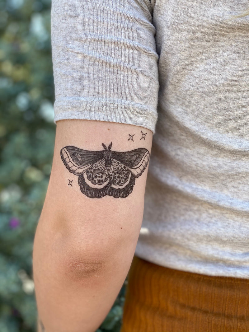 Temporary tattoo of a night moth with details and sparkles on a person's forearm wearing white and orange with a blurred nature background 