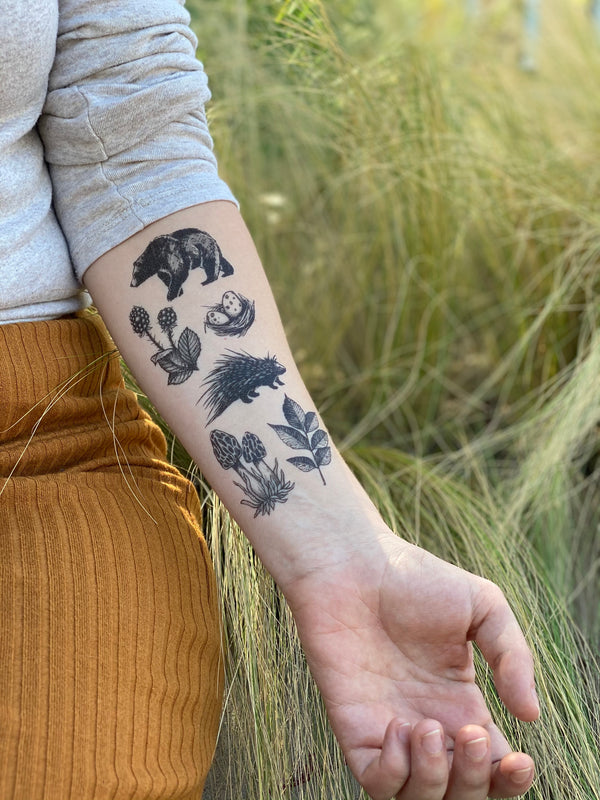 A person's forearm displaying a collection of woodsy temporary tattoos, including images of a bear, porcupine, berries, and mushrooms.
