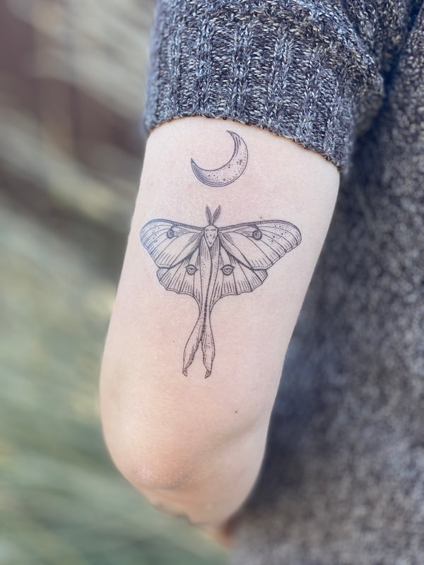 Luna Moth with crescent moon above temporary tattoo on back of persons upper arm wearing a grey sweater in a blurred nature background 
