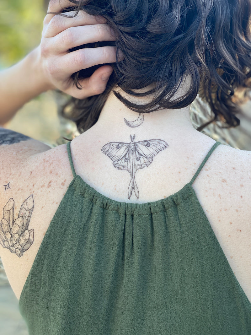 Luna Moth with a crescent moon above it temporary tattoo on the back of persons neck wearing green in a blurred nature background 