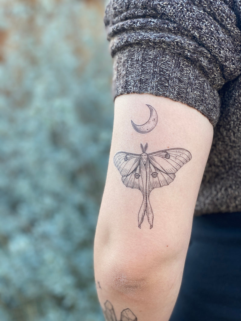 Luna Moth with crescent moon above it temporary tattoo on persons arm wearing blue and grey in a blurred nature background 