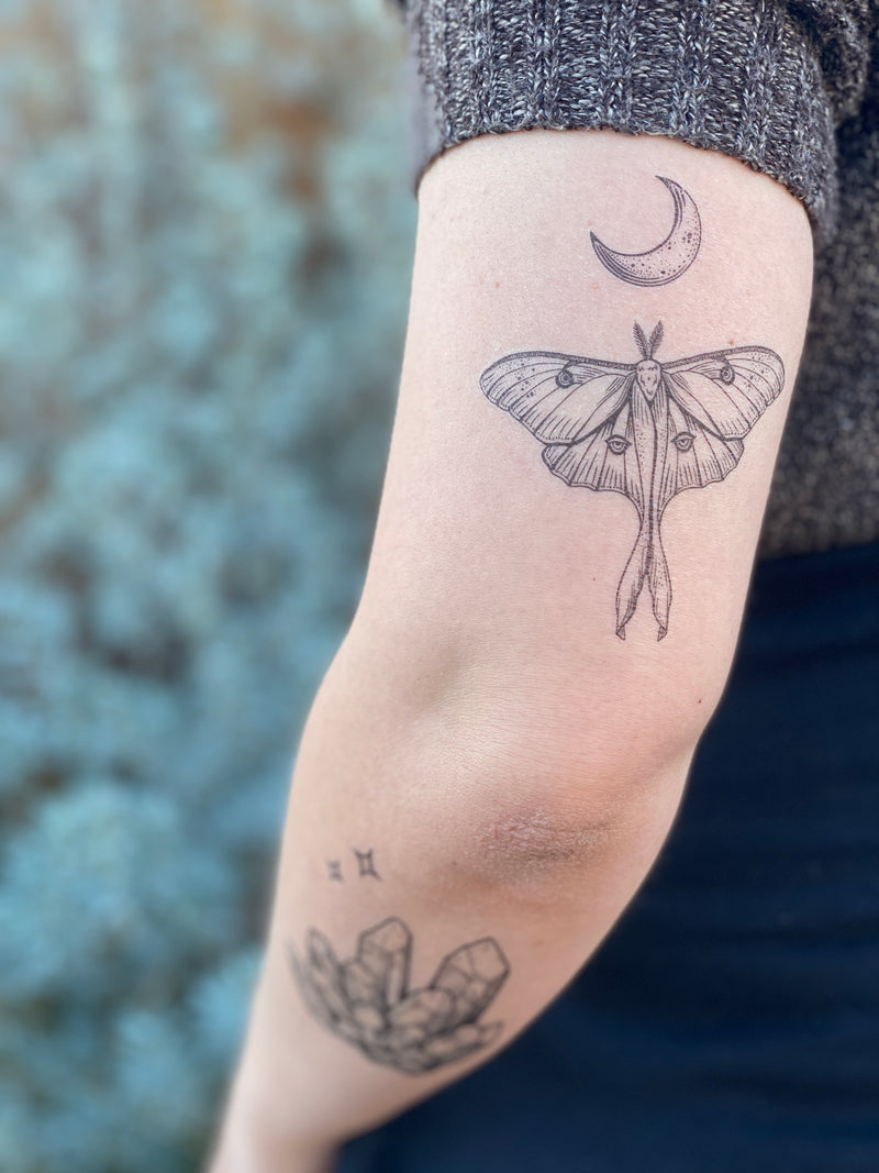 Luna Moth with crescent moon above it temporary tattoo on persons arm wearing blue and grey with a blurred nature background 