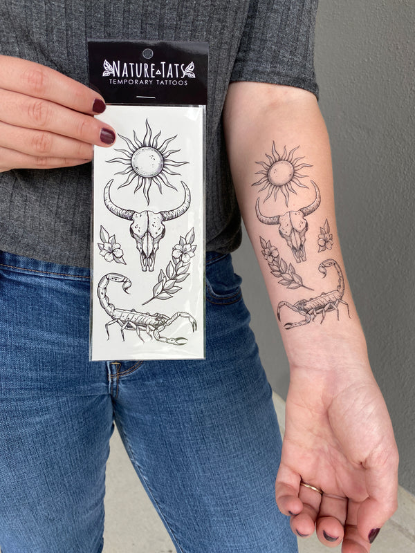 sunlit south temporary tattoo collection on persons arm wearing grey and blue holding a temporary tattoo in it packaging, with a grey background 